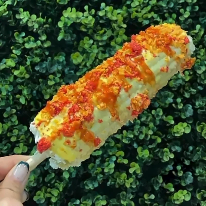 Elote entero preparado con queso y chile en polvo, sostenido frente a un muro de hojas